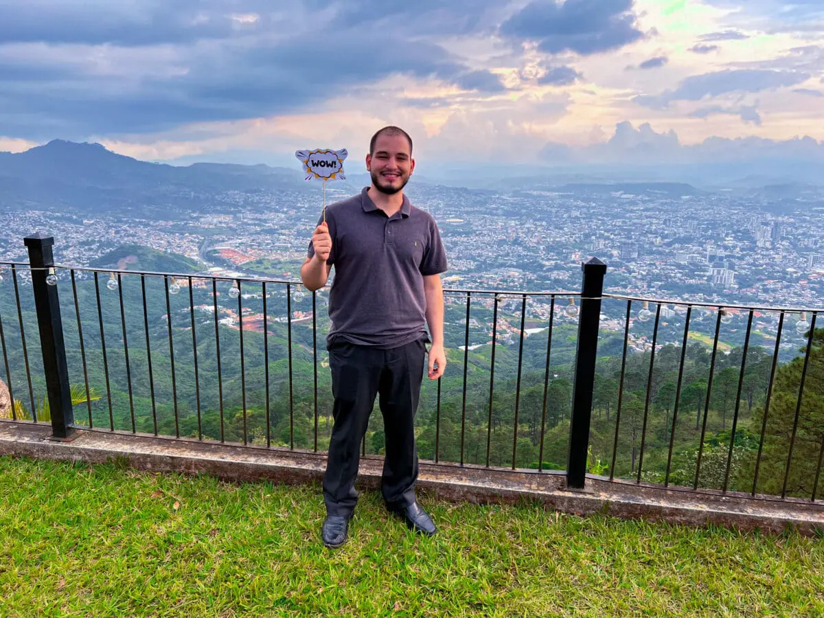 Miembro del equipo de Andes Development sonriendo con un letrero de 'WOW!' durante el evento de celebración corporativa por los 12 años de la agencia, con un hermoso atardecer y la ciudad de fondo.