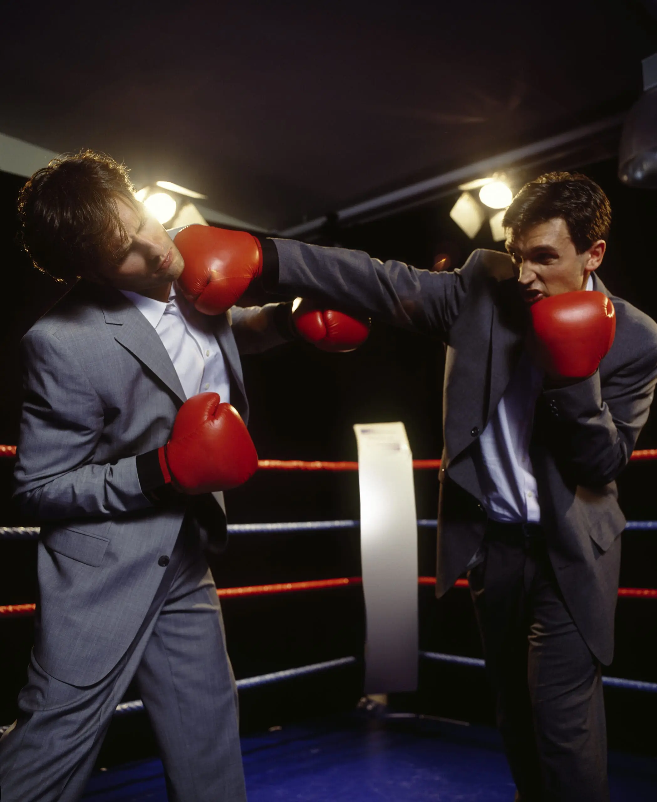 Dos hombres de negocios con trajes grises y guantes de boxeo rojos peleando en un ring. Representa de forma metafórica el conflicto y debate tradicional sobre qué lenguaje de programación elegir, un problema que se soluciona con el enfoque Dual Stack (Python y Laravel).