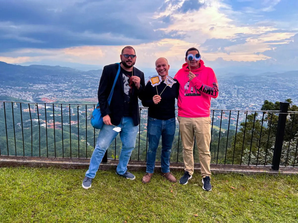 Tres colaboradores de Andes Development compartiendo un momento ameno con accesorios festivos durante la celebración de los 12 años de la empresa. La fotografía al aire libre muestra una espectacular vista de la ciudad y las montañas de fondo, reflejando el compañerismo en la agencia.
