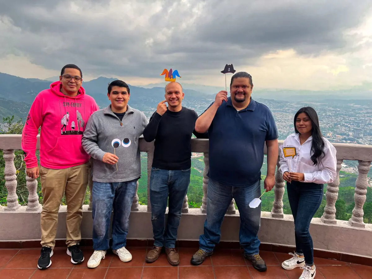 Grupo de cinco integrantes del equipo de Andes Development compartiendo un momento agradable con divertidos letreros de utilería durante la celebración de los 12 años de trayectoria de la agencia. Fondo de paisaje urbano al atardecer.